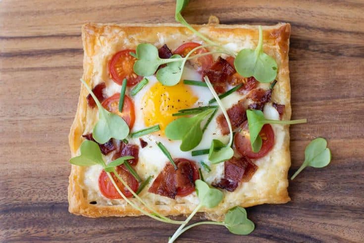 A square of puff pastry topped with cheese, tomatoes, bacon, egg, chives and greens