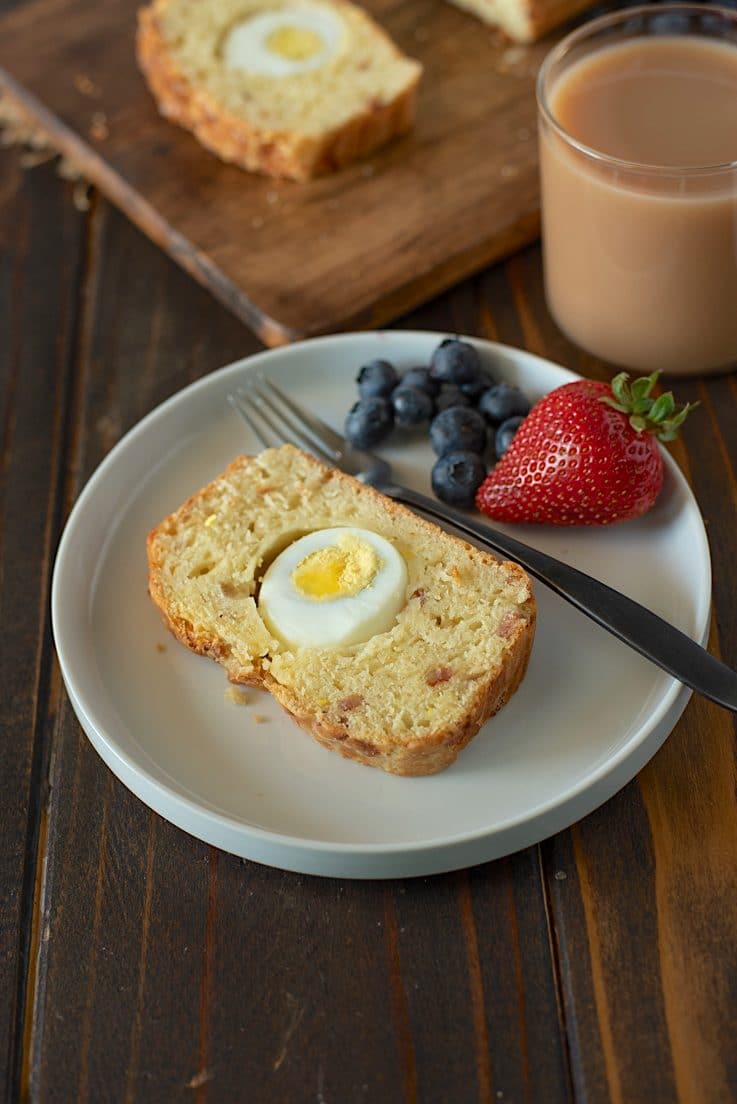A slice of loaf on a white plate with a fork, strawberries, blueberries and a cup of coffee