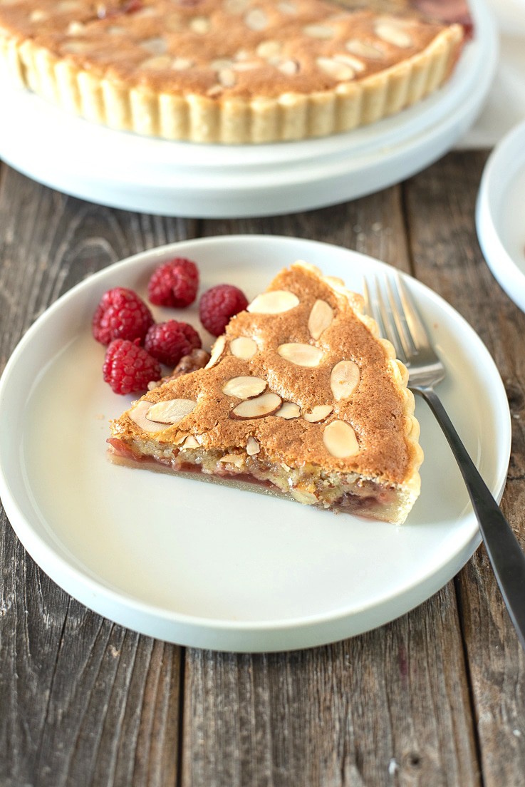 A slice of bakewell tart on a place showing the strawberry jam layer with a fork and raspberries