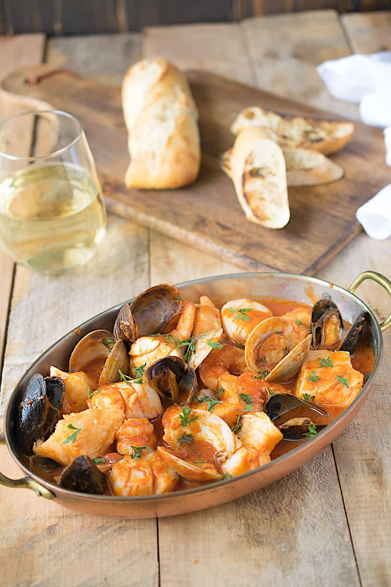Fish and shellfish in an oval serving dish with a glass of wine and crusty bread
