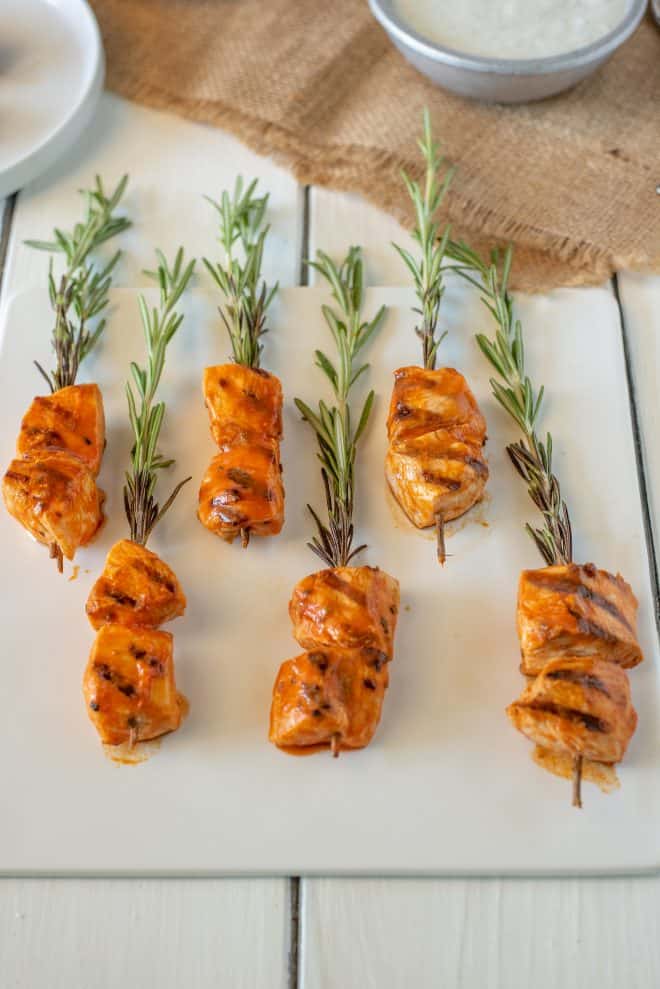 Pieces of chicken coated in buffalo sauce skewered onto fresh rosemary sprigs lying on a square white plate