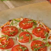 7 slices of tomato melted cheese and fresh basil ribbons top a cauliflower crust pizza with fresh basil and tomatoes in the background