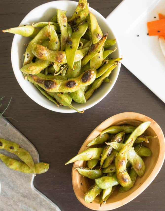 A white bowl and a wooden bowl filled with charred edamame with some spilling out