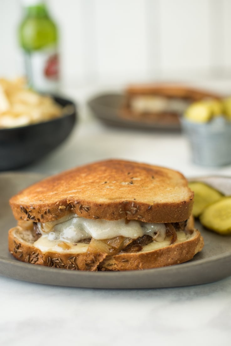 A patty melt sandwich on a plate before being cut showing delicious melted cheese oozing out the side