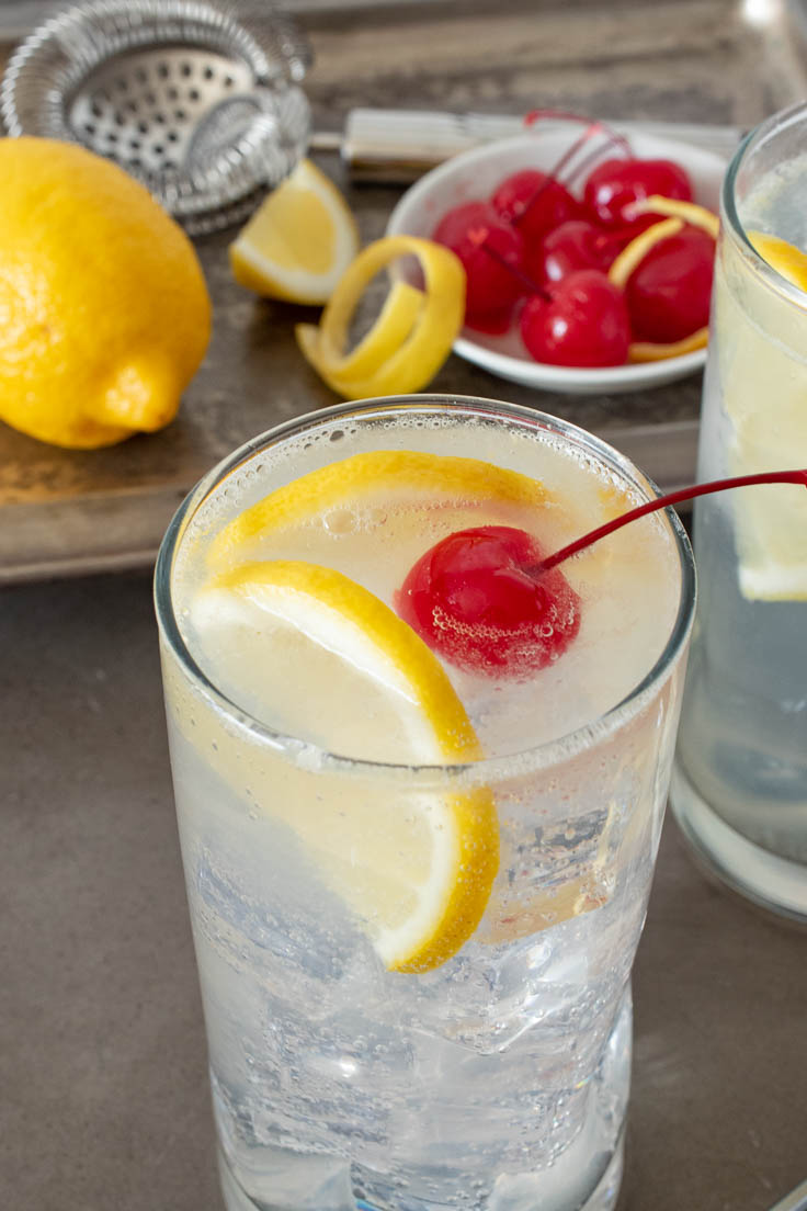 Lemon slices and red cherry in a glass of Tom Collins with lemons and cherries on a serving tray