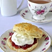 A scone cut in half topped with jam and clotted cream with a flower decorated china tea cup filled with tea