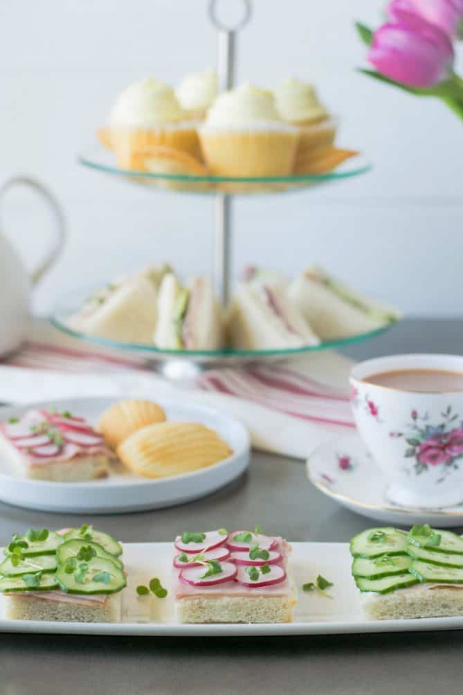 A selection of cucumber and radish open face sandwiches with a cup of tea, a tiered cake stand with pastries and sandwiches