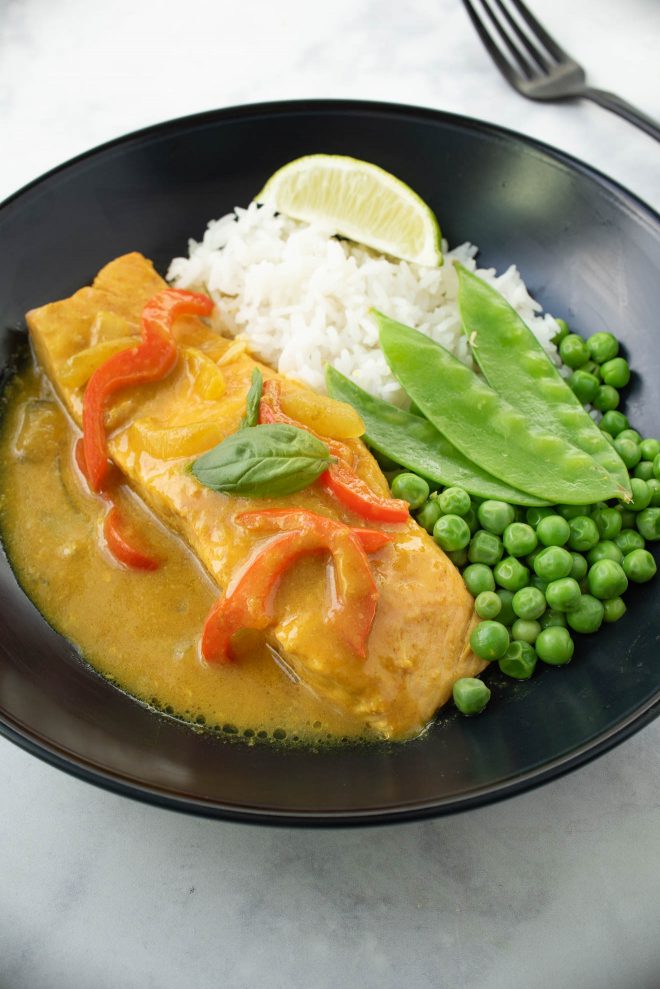 A complete meal in a black bowl, salmon in curry sauce with peas, slow peas and white rice.