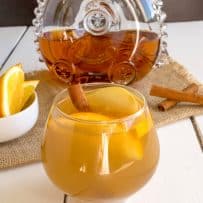 A short glass of Cognac apple cider garnished with orange wedges, apple wedges and a cinnamon stick