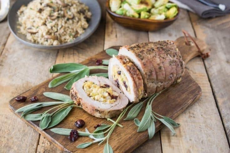 Apple cranberry cornbread stuffed pork loin served on a cutting board garnished with fresh sage leaves with a bowl of Brussels sprouts and rice in background