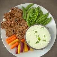 Tzatziki dipping sauce in a grey bowl on a white plate surrounded by vegetables and crackers.