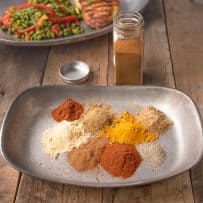 A plate with piles of different color spices with a plate of grilled chicken and peas in the background