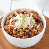 A white bowl full of turkey and bean chili topped with cheddar cheese and cilantro