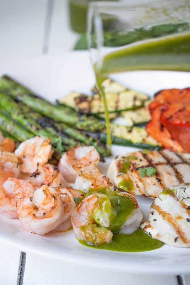 Pouring sauce over grilled shrimp, chicken and vegetables on a white platter