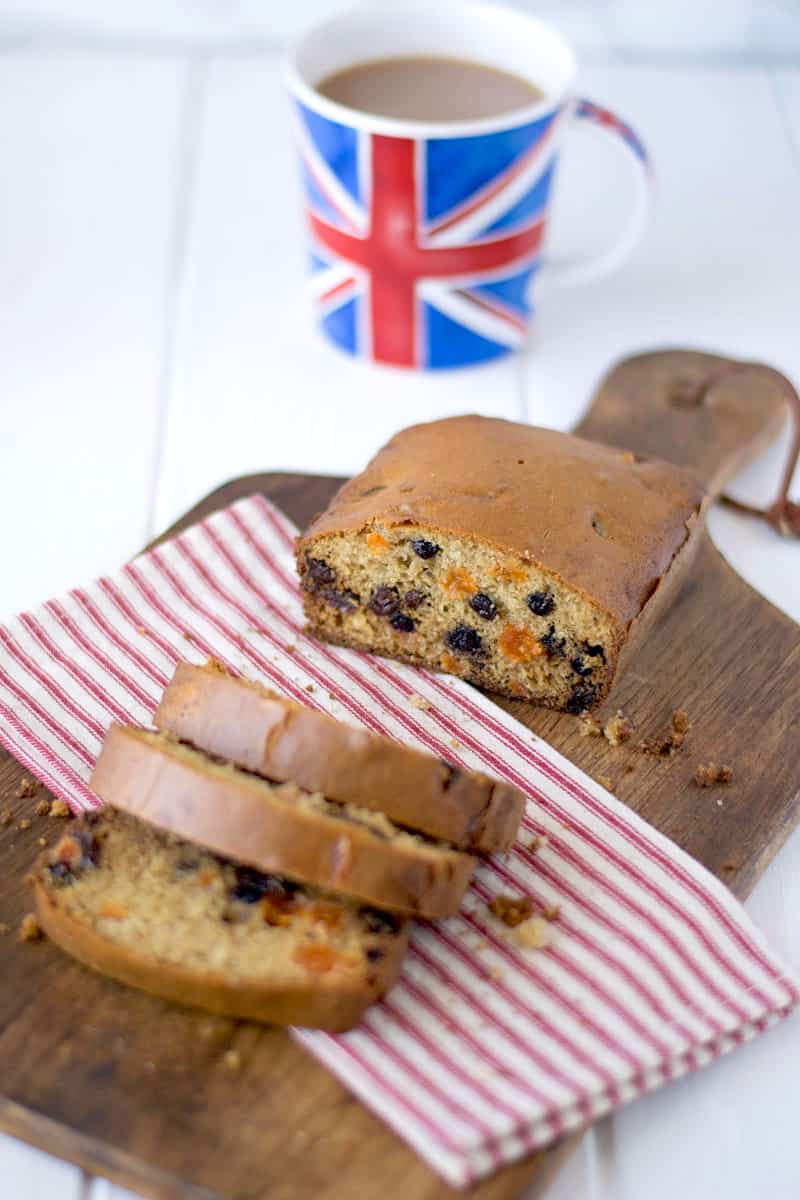 Tea cake studded with fruit on a cutting board, sliced with a cup of tea