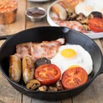 Fried egg, bacon, sausage, tomato, mushrooms in a cast iron pan.