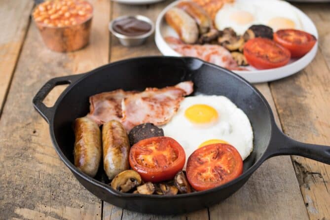 Fried egg, bacon, sausage, tomato, mushrooms in a cast iron pan.