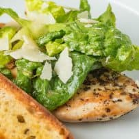 A chicken breast peeking out from under romaine lettuce topped with shaved Parmesan and a crouton