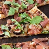 A slice of grilled flatbread topped with fontina, sliced fresh figs, prosciutto, arugula leaves and balsamic glaze