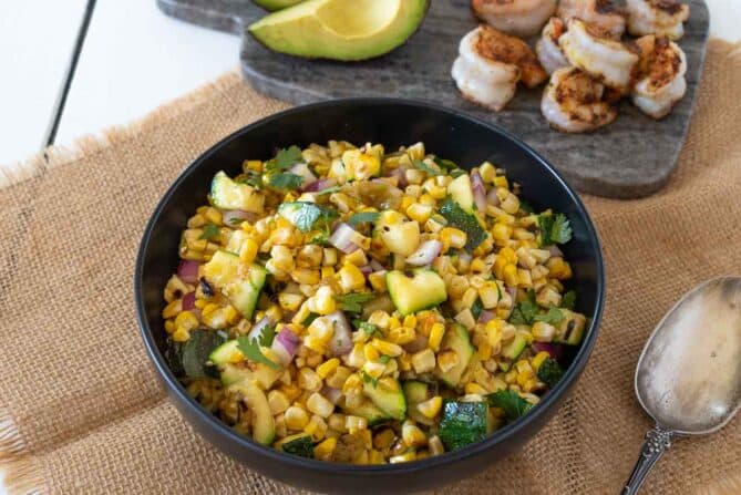 A black bowl filled with corn kernels and zucchini with a serving spoon and grilled shrimp