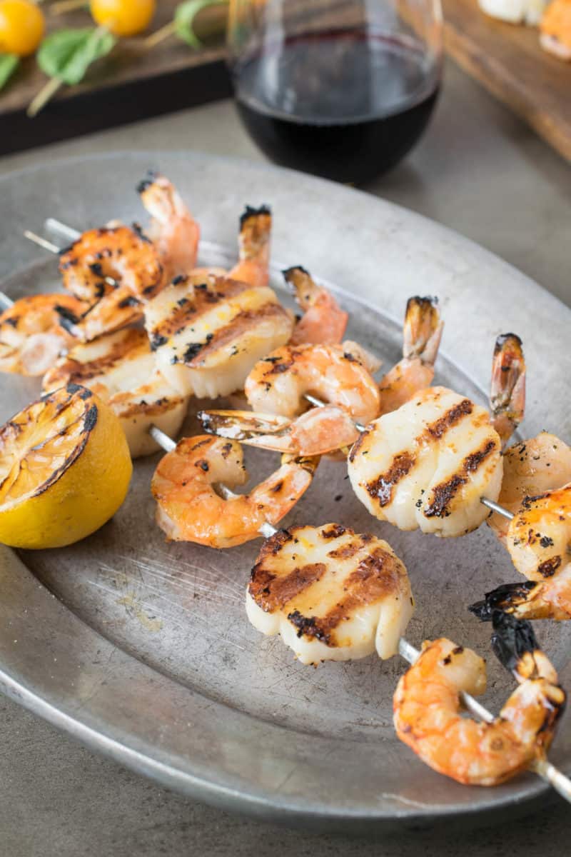 Grilled shrimp and scallops on skewers on a pewter plate with fresh lemon half and a glass of wine