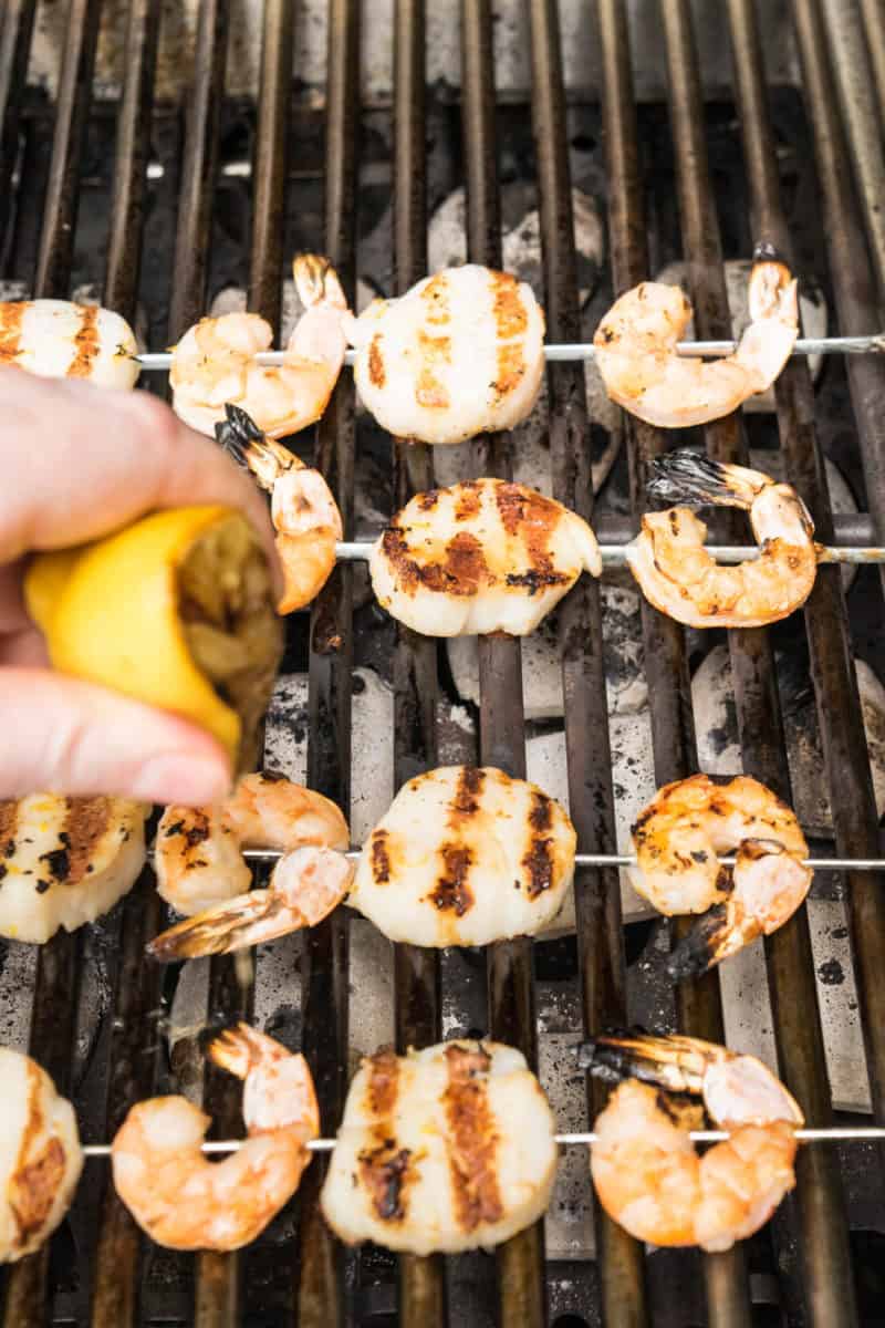 Skewered shrimp and scallops on a grill with fresh lemon being squeezed onto them