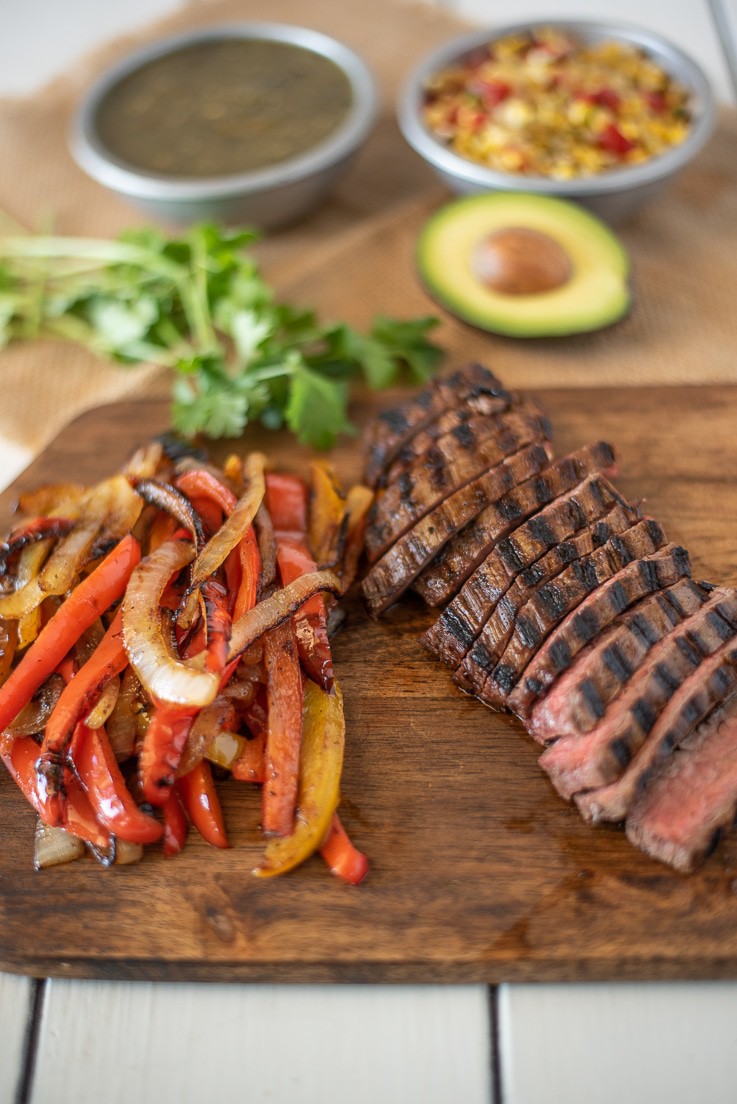 Sliced, grilled steak and grilled peppers and onions on a board with avocado and salsa ready to build the bowl
