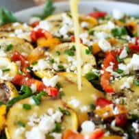 Pouring dressing over a platter of grilled squash and peppers and feta