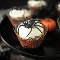 A closeup of a cupcake in a spider web muffin cup topped with a large plastic spider with mini orange pumpkins around