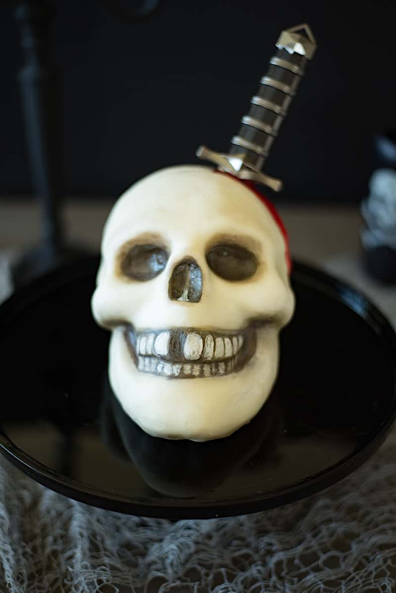 A skull cake covered in white fondant and painted with food paint with a real dagger and red blood