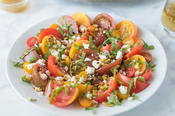 Multi-color sliced tomatoes on a round white plate topped with toasted pistachios, feta and fresh basil