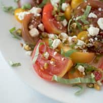 A closeup of a juicy, red and yellow heirloom tomato with chopped basil, feta and pistachios