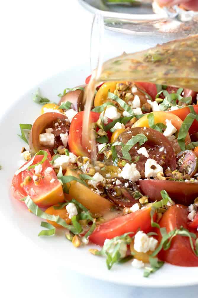Pouring dressing onto a platter of sliced tomatoes with pistachios, feta and fresh basil