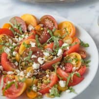 Slices of different color heirloom tomatoes, large and small on a plate topped with crumbled feta cheese, toasted pistachios and basil