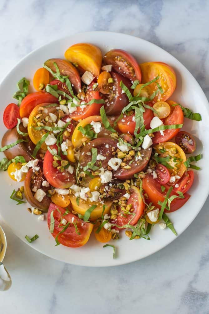 An overhead view of the yellow, orange and red tomatoes on a round plate with feta cheese and ribbons of basil