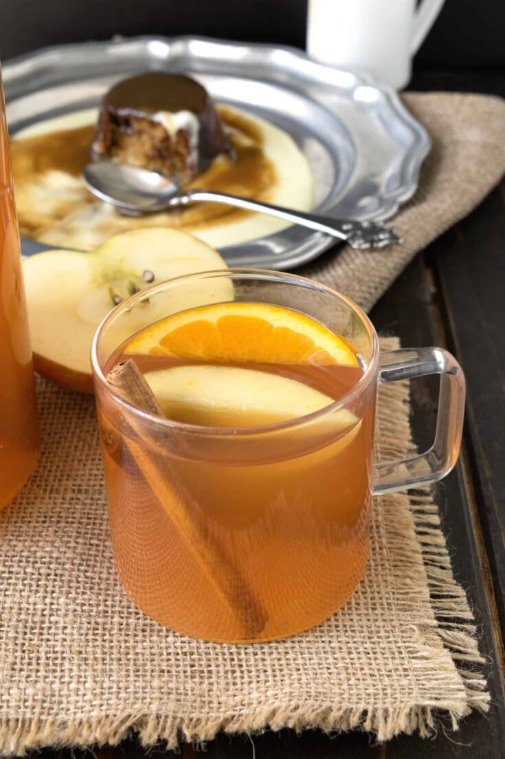 A glass mug of apple cider garnished with an apple wedge, orange slice and cinnamon stick