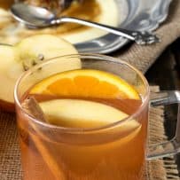 A golden colored hot apple cider in a glass mug with apple, orange and cinnamon stick