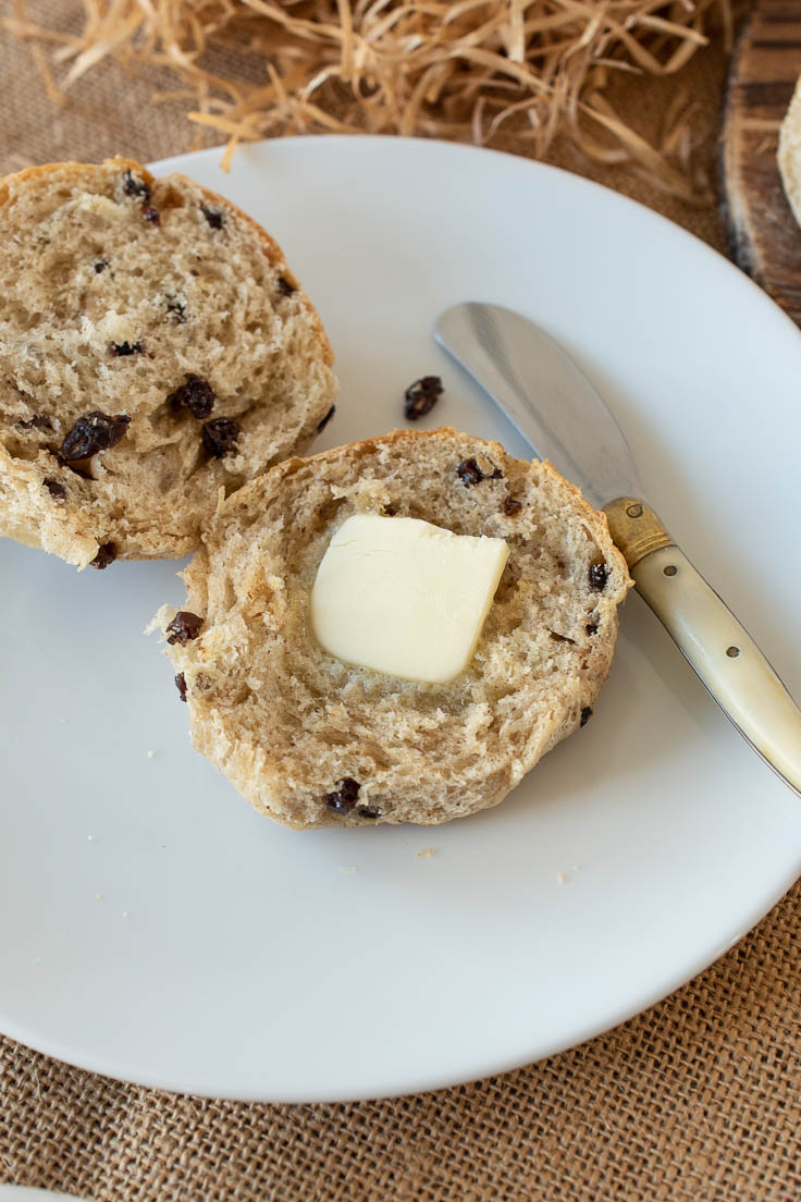 A hot cross bun cut open on a plate with butter and a knife
