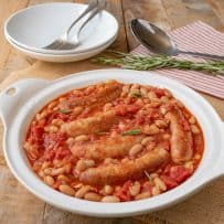 A round white serving bowl with sausage and beans with 2 bowls and forks