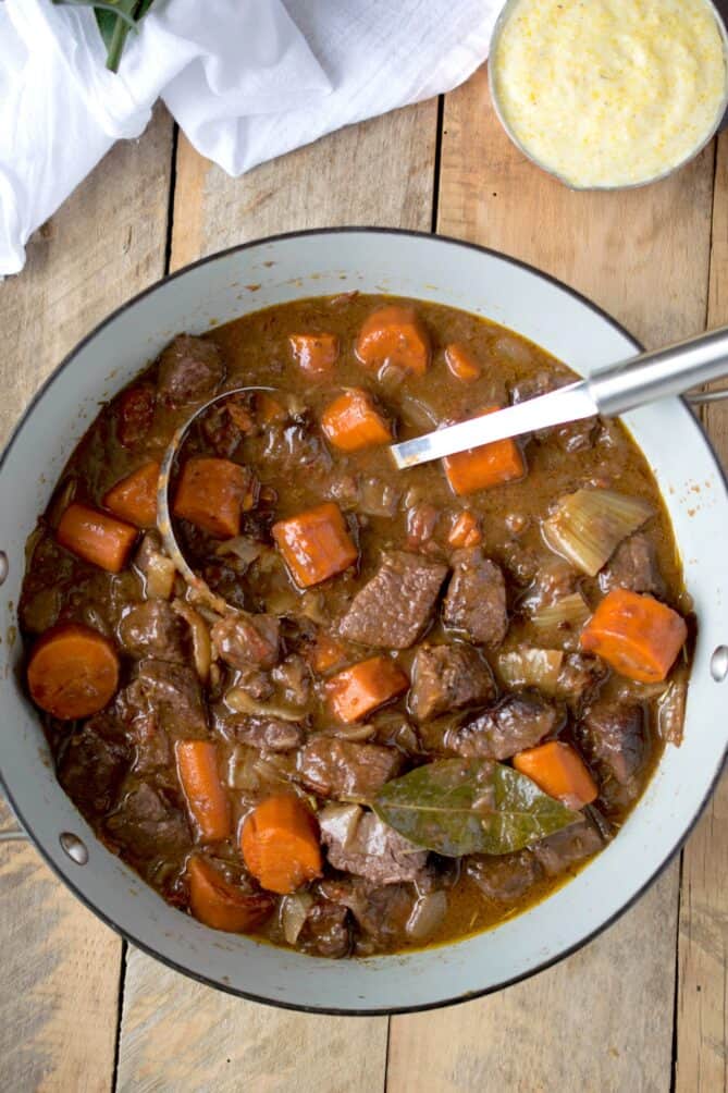 A large pan of beef stew with beef, carrots and a bay leaf with a ladle
