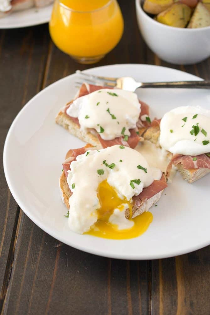 A poached egg on top of prosciutto and an English muffin cut open with yellow yolk running out