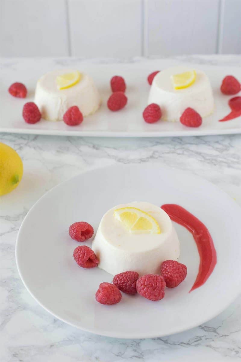 Lemon panna cotta on a white plate surrounded by raspberry sauce and fresh raspberries