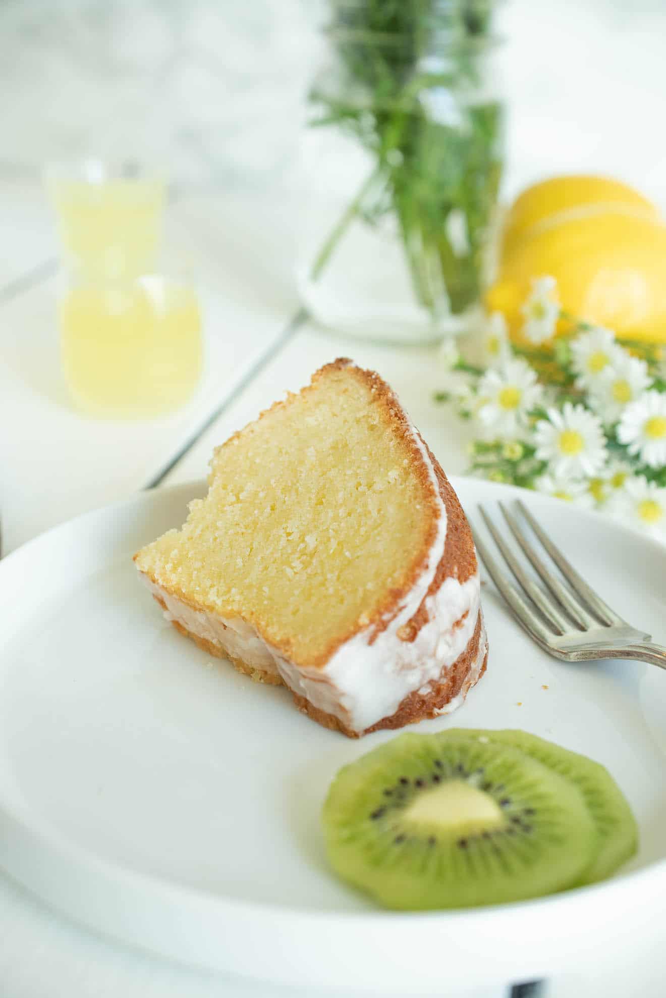 A slice of limoncello pound cake on a white plate with a fork and slices of kiwi