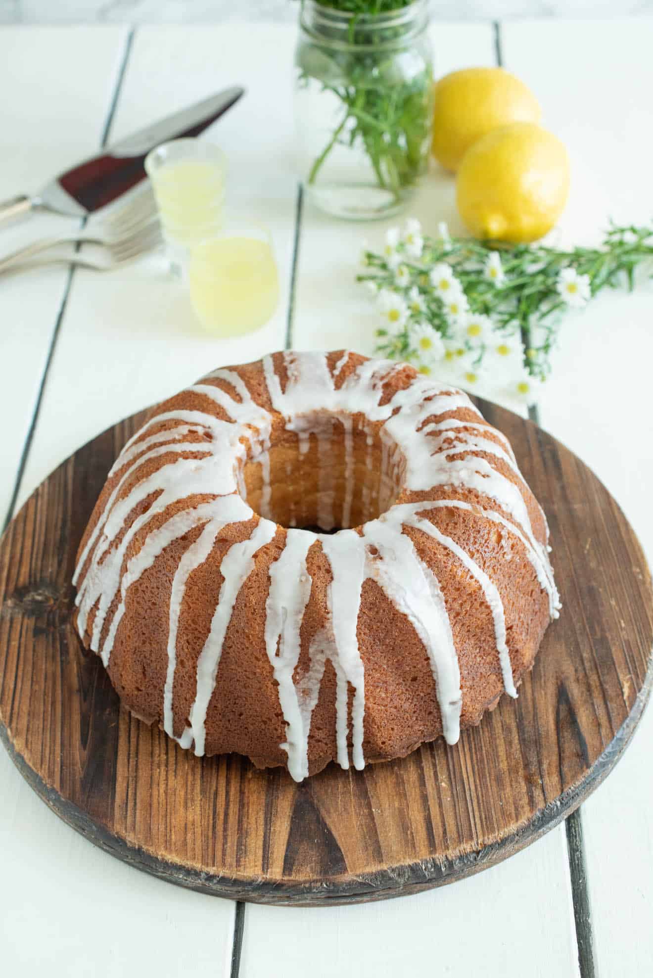 A whole bundt cake drizzled with frosting on a round, wood cake stand with lemons and daisies in the background