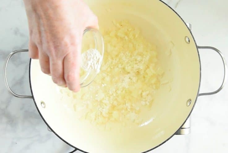 All purpose flour being sprinkled into the pan of onions and garlic