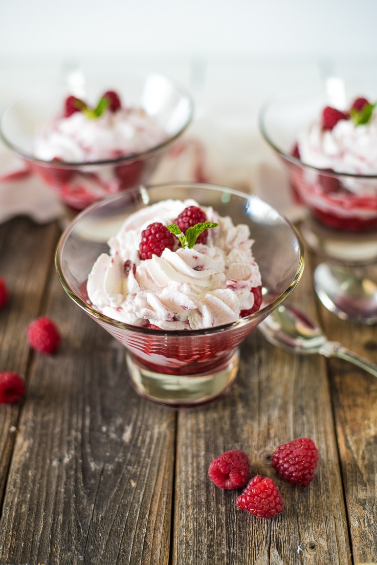A glass bowl with raspberry fruit fool surrounded by fresh raspberries and spoons