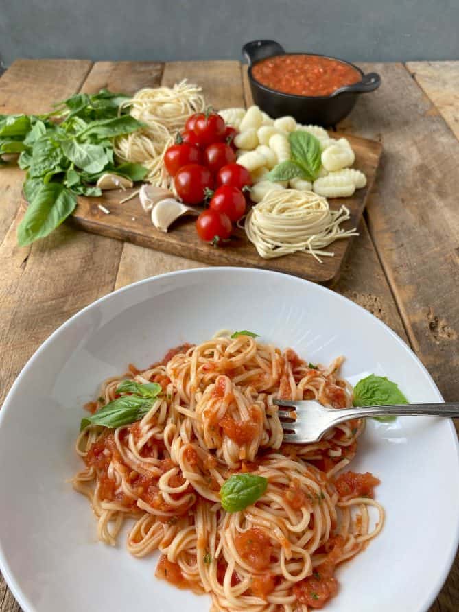 A bowl of pasta with fresh tomato sauce with a board of fresh pastas, tomatoes and basil behind