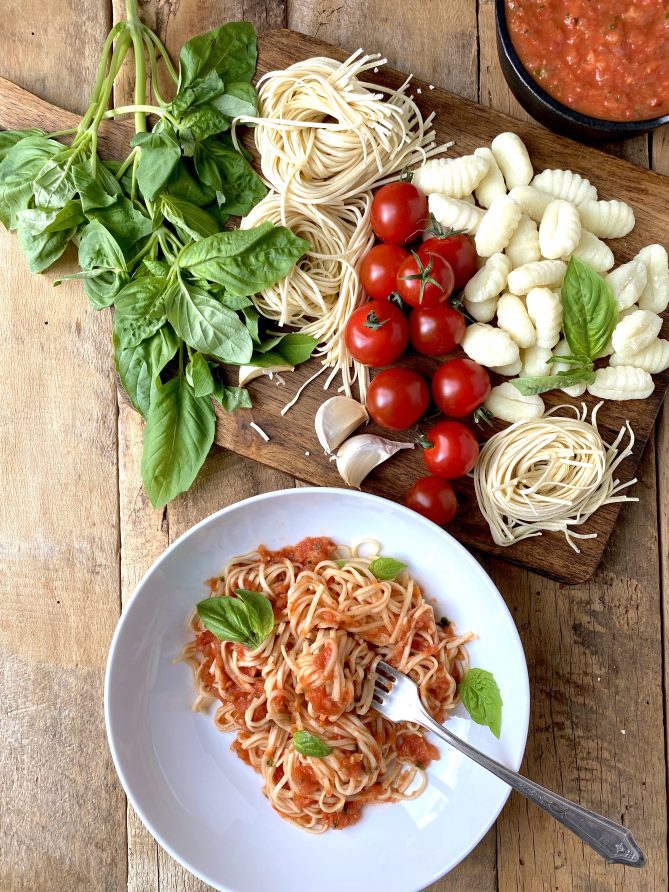 A display of fresh basil leaves, pasta and tomatoes viewed from overhead with a bowl of pasta and sauce