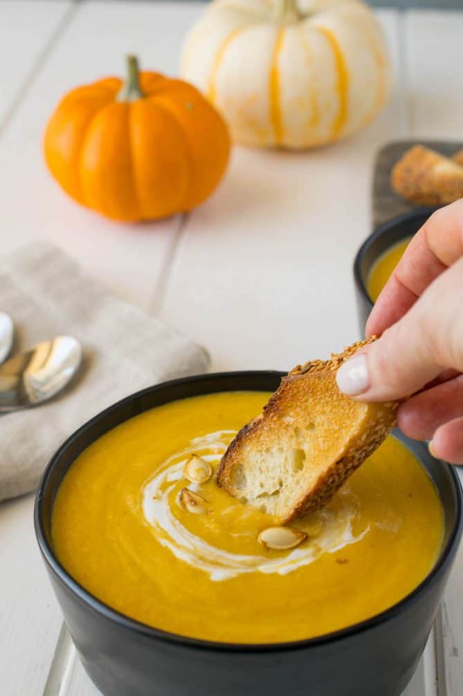 Dipping a piece of toasted bread into the pumpkin and roasted root vegetable soup with mini pumpkins in the background