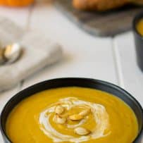 Smooth, mustard color roasted pumpkin and root vegetable soup in a black bowl topped with sour cream and toasted pumpkin seeds
